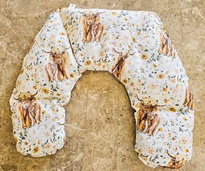 Hot and Cold Neck Pack with a white background, featuring a print of Highland cows and yellow flowers, laid flat on a textured beige stone surface.