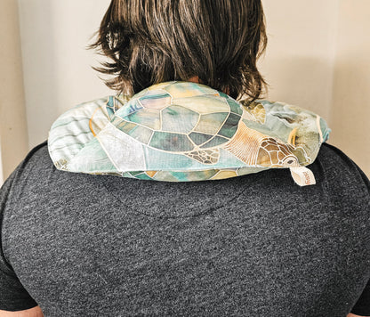 A person viewed from behind wearing a turtle-patterned hot and cold neck pack on their shoulders. The neck pack features a mosaic design in shades of teal, green, beige, and brown, with a small white fabric tag visible on the right side.