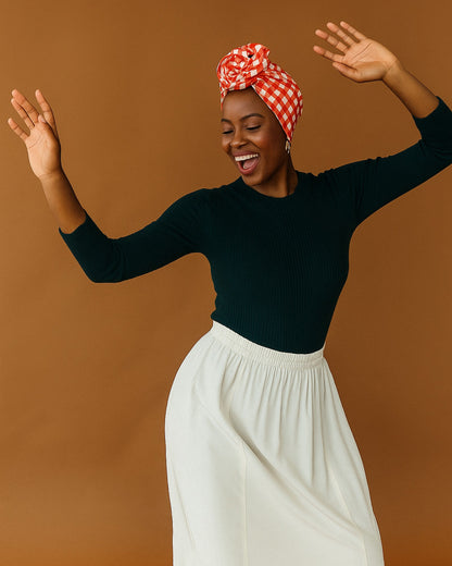 Woman wearing a black top and white pants with a red and white checkered wire headwrap against a brown background