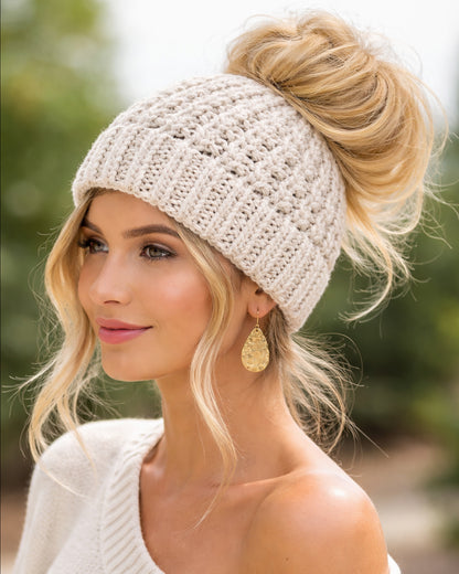 Woman wearing a beige knit beanie with her hair styled in a bun, outdoors.